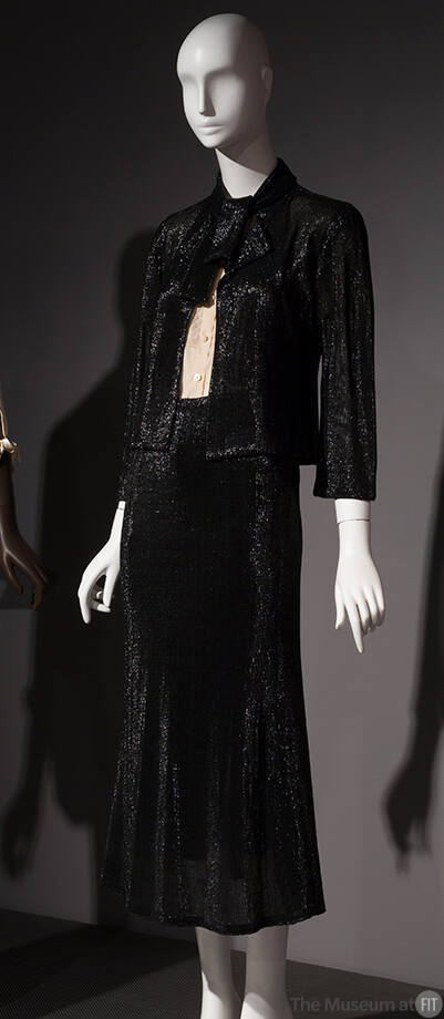 Black suit with fitted, tie collar jacket, calf length A-line skirt and white button front shir…