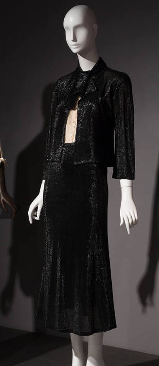 Black suit with fitted, tie collar jacket, calf length A-line skirt and white button front shir…
