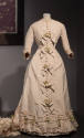 Ivory dress with round neck, self bows and artificial flowers down front, pleated trim at hem
