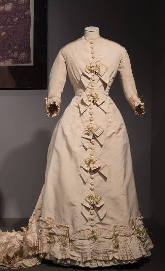 Ivory dress with round neck, self bows and artificial flowers down front, pleated trim at hem