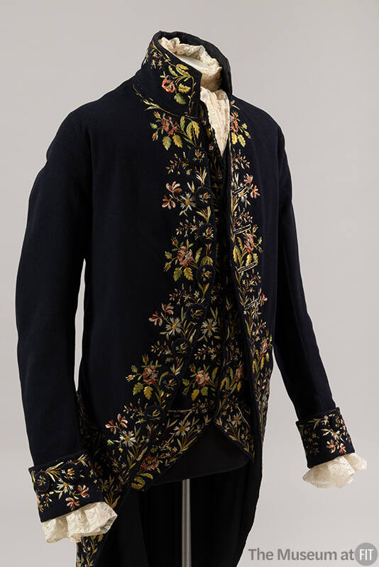 Man’s navy blue coat and matching vest with multicolor floral trim and stand collar, dressed wi…