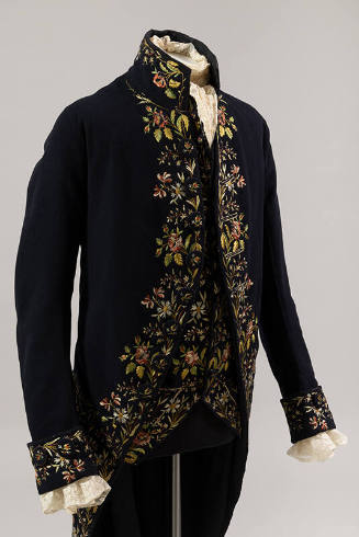 Man’s navy blue coat and matching vest with multicolor floral trim and stand collar, dressed wi…