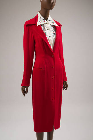 ¾ view of knee length red fitted coat dress with jacket styling, long stylized collar points an…