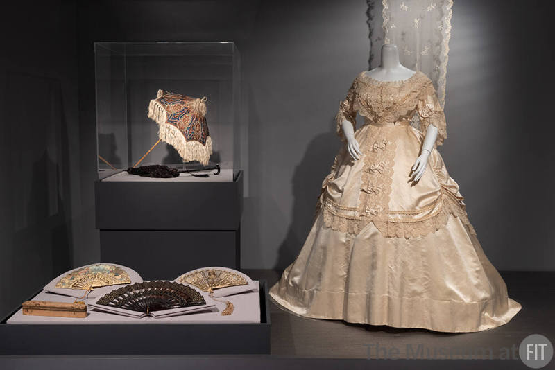 Silk evening gown, circa 1869, with lace shawl on wall, circa 1850, fans by J. Duvelleroy and unidentified designers, circa 1840-circa 1860, and parasols, 1840-1870