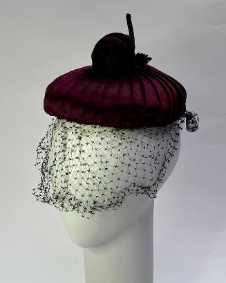 Small circular burgundy velvet hat topped with velvet rose; net face veil 
