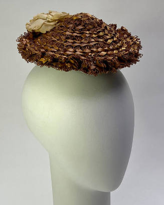 Small light brown toque hat made of loops of threads with ribbon woven through in concentric ri…