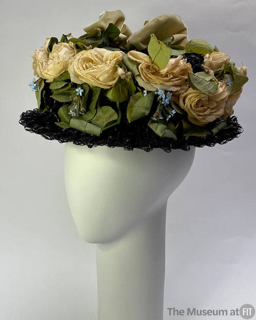 Black straw hat with off-white roses, green leaves and tiny blue flowers covering crown and lig…