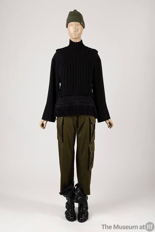 Black cable knit vest over long sleeve black top with hem fringe, green cargo pants, black leat…
