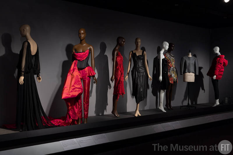 Dior + Balenciaga, Left to right: 2007.46.39 (black evening dress), 99.83.9 (corset evening ensemble), 2021.53.34 (red dress), 2001.45.1(black leather dress), 2008.60.1 (black one shoulder dress), 2005.23.1 (graffiti knit dress), 2019.21.1 (jacket), 2016.113.1 (red puffer coat)