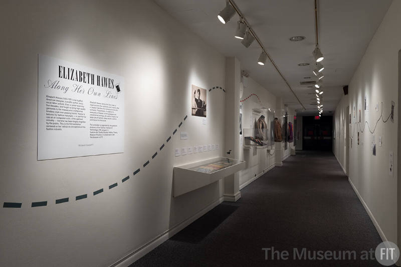 Elizabeth Hawes exhibition installation view of a dimly lit hallway with wall-mounted cases of …