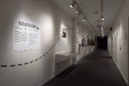Elizabeth Hawes exhibition installation view of a dimly lit hallway with wall-mounted cases of …