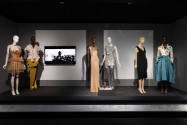¡Moda Hoy! exhibition installation view of six dressed mannequins, a pair of shoes in a case an…