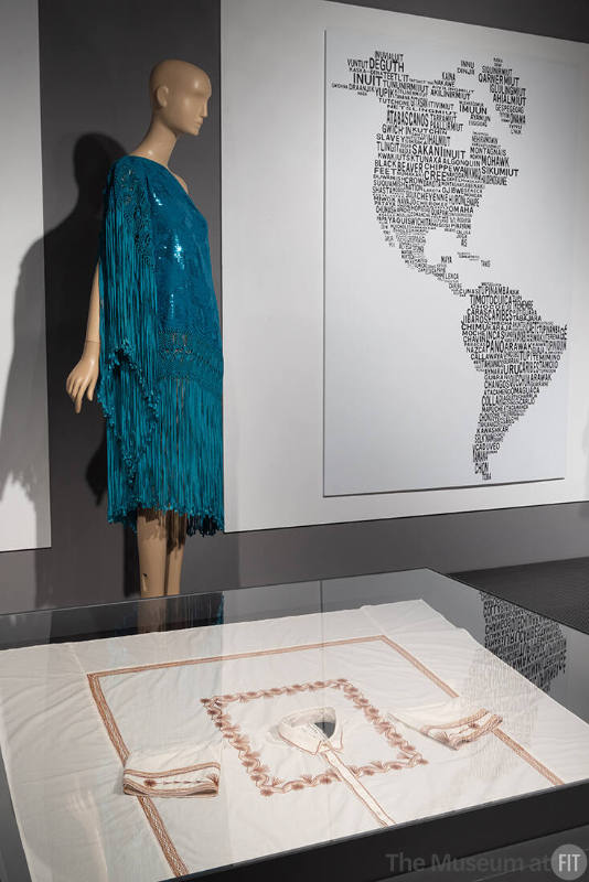 Installation view featuring the Chola Asymmetrical dress by the Bolivian Fashion brand  Juan de la Paz and artisan Julia Mayta (2022.81.1); Cuadro Tlahui blouse (2022.26.1), a collaboration between Guillermo Vargas of 1/8 Takamura and Paula Pérez Vázquez (Mixe) of Artesanía Textil Arte-Fer and reproduction of the Original Peoples of the Continent artwork by Mexican fashion designer Carla Fernández and artist Pedro Reyes.