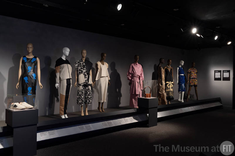 View of the “Sustainability” and “Craftsmanship” sections in the exhibition ¡Moda Hoy! Latin American and Latinx Fashion Design Today. From left to right: 2023.5.1 (shoes case), 2023.8.1 (black dress), 2022.50.2 (set), 2022.50.1 (set), 2023.2.5 and 2023.9.1 (top and pants), 2019.10.1 (pink set), 2023.12.1 (bag case), 2010.48.1 (tan gown), 2022.69.1 (black dress), 2015.27.1 (blue dress), 2007.49.1 (beaded jumpsuit)