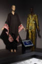 ¡Moda Hoy! exhibition installation view of two mannequins in a black cape and a yellow, blue an…