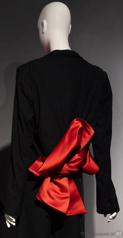 Back view of black jacket with slightly flared sleeves and large red/orange bow at waist