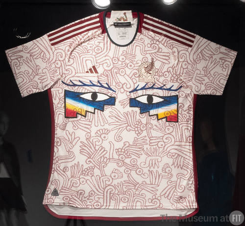 Off-white short sleeve athletic jersey with allover red print of Mixtec and Aztec deities and b…