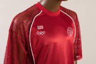 Detail of red uniform jersey top, net sleeves, white Olympic rings and white Telfar logos on ch…