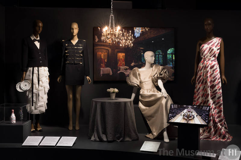 Food & Fashion Left to right: Judith Leiber Couture, Ice Champagne Bottle Bubbly minaudière, Fall 2021. Lent by Judith Leiber Couture; 2015.64.1 (waiter ensemble), 2020.6.1 (black suit), 88.105.10 (beige dress), 2009.19.1 (white printed dress)