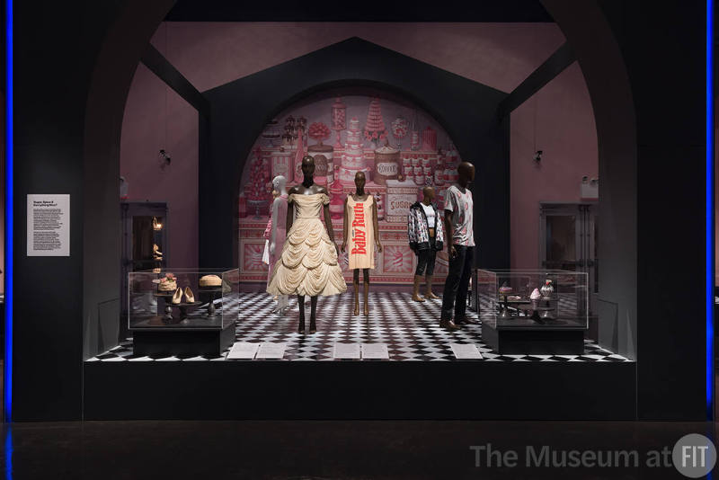 (Left) Balenciaga, hat, c.1955; shoes, c.1932; Bergdorf Goodman, hat with pink bow, c.1965. (Center) Jean Dessès, cream dress, c.1957; Mars of Asheville, paper dress, c.1968. (Right) Judith Leiber Couture, donut Strawberry Sprinkle minaudière, fall 2019, ice Cream Sundae Sprinkles minaudière, pre-fall 2019, ice Cream Cone Tutti Frutti minaudière, fall 2020, Lent by Judith Leiber Couture. ICECREAM, popsicle print T-shirt, c.2010; Ricardo Seco, Immigrant Flavors ensemble, fall 2021, Lent by Ricardo Seco. Background image: Reproduction of Mark Ryden, Dessert Counter, 2016, Courtesy of the artist and Kasmin Gallery.