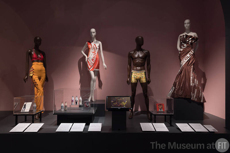 CHROMAT, red and orange ensemble with Cheetos bag, fall 2018; (case) Karl Lagerfeld, The Karl Lagerfeld Diet book, 2005; Karl Lagerfeld, limited-edition Coke Light bottle, 2011, Lent by Melissa Marra-Alvarez. Jeremy Scott for Moschino, “Drink Moschino” cola can print dress, stretch rayon, resort 2015, The Museum at FIT; (case) Coca-Cola, Limited edition-bottles designed by Jean Paul Gaultier, 2012; Diane von Furstenberg, 2012; Marc Jacobs, 2013, Lent by Melissa Marra-Alvarez; (case) Candy's Bikini, candy string bikini, 1982;  Versace, fruit, butterfly, and floral print swim trunks, 1990s; Jeremy Scott for Moschino, chocolate bar dress, fall 2014.