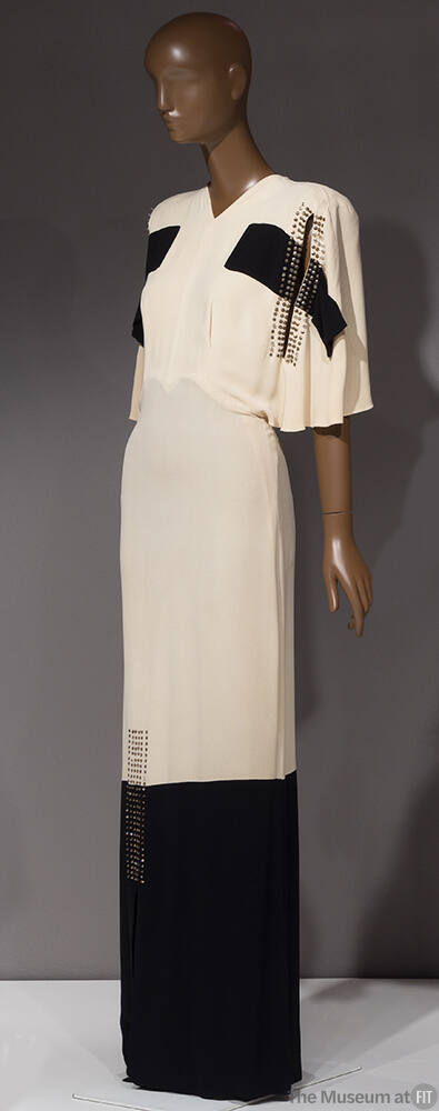 White floor length evening dress with cape-like elbow length sleeves, black bands across either…