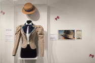 Untying the Bow exhibition installation view of a beige jacket and straw boater hat in a case n…