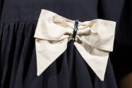 Detail view of navy blue tent dress showing ivory bow with oblong ornament of multicolor glass …