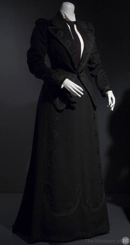 Black suit trimmed with loops of black braid on lapels, cuffs and front panel of floor length f…
