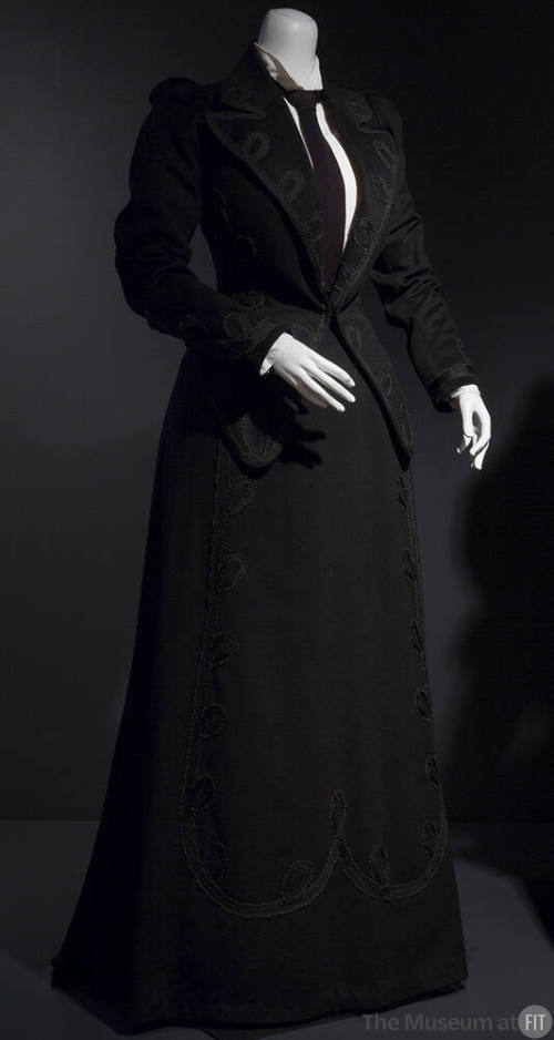Black suit trimmed with loops of black braid on lapels, cuffs and front panel of floor length f…