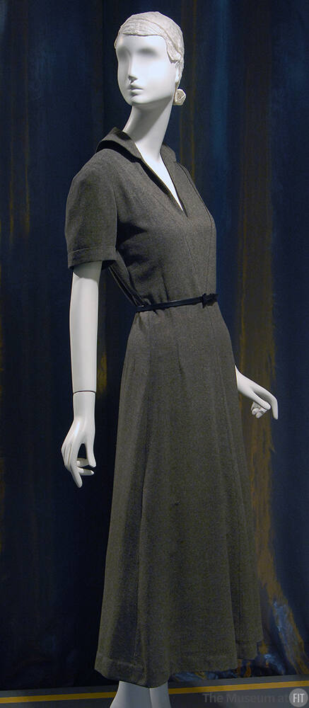 ¾ view of dark grey short sleeve ankle length dress with rounded collar, deep V-neck and slim b…