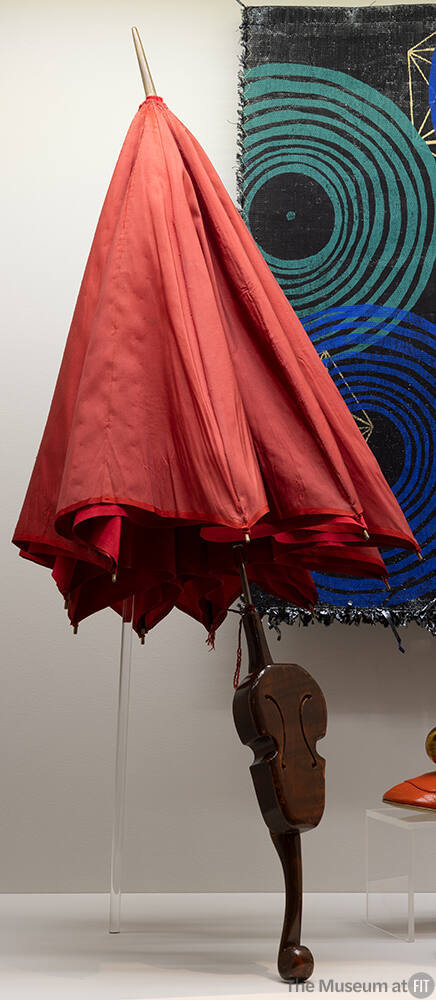 Red umbrella with miniature violin in center of wooden handle shown in front of a black textile…
