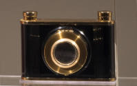 Cosmetic case in the shape of a black camera with gold trim on top and bottom and around the le…