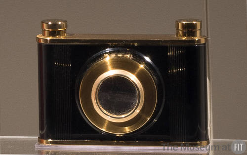 Cosmetic case in the shape of a black camera with gold trim on top and bottom and around the le…