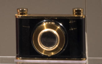 Cosmetic case in the shape of a black camera with gold trim on top and bottom and around the le…