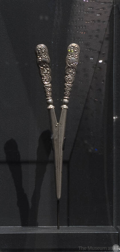Silver glove stretcher with two tapering scroll patterned handles that meet at hinge and extend…