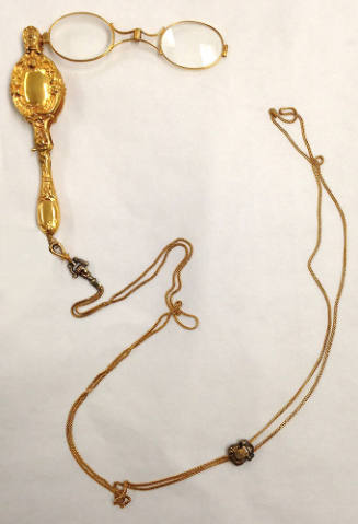 Gold lorgnette of eyeglasses with small oval frames attached to collapsible case with scrolling…