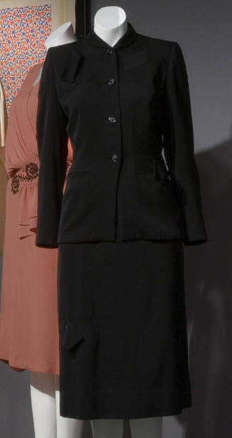 Black suit with high shawl collar, four button center closure, tabs on left shoulder and right …