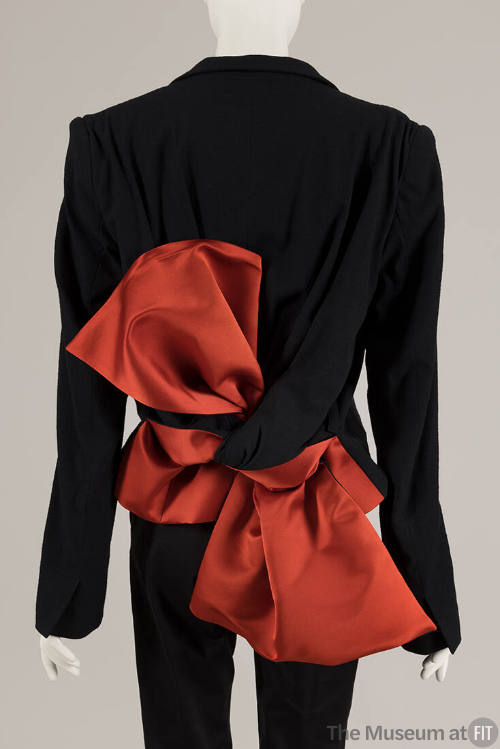 Back view of black jacket with tails extending from shoulders and tied into oversize red/orange…
