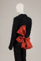 Back ¾ view of black jacket and pants with oversize red/orange bow tied at waist