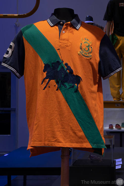 Man’s orange polo shirt with navy blue short sleeves and collar, a diagonal green stripe and la…