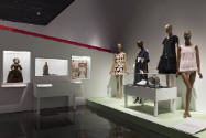 Doll Dressing exhibition installation view of several wall cases with dolls and a platform with…