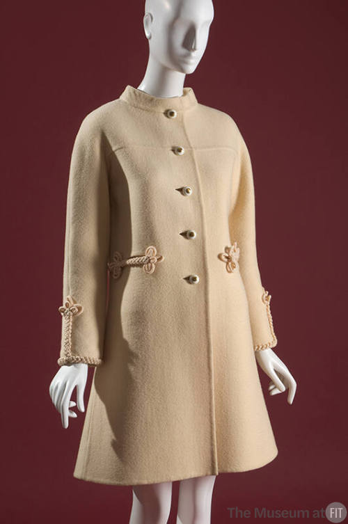 Cream thigh-length coat with short collar, metal buttons at center front and braided details at…