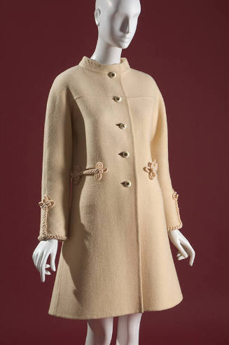 Cream thigh-length coat with short collar, metal buttons at center front and braided details at…