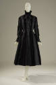 Black coat with a high collar with bow, long sleeves with pointed cuff, pocket over hips, and f…