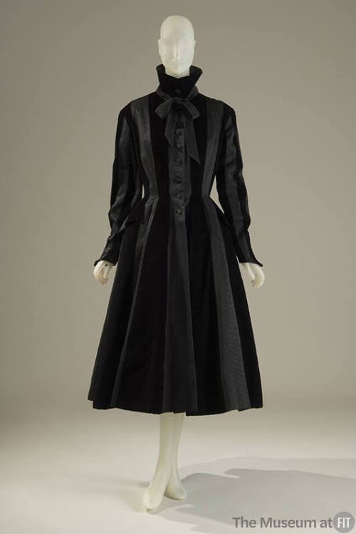 Black coat with a high collar with bow, long sleeves with pointed cuff, pocket over hips, and f…
