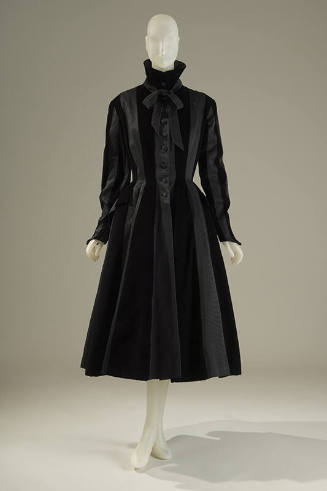 Black coat with a high collar with bow, long sleeves with pointed cuff, pocket over hips, and f…