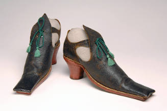 Front & 3/4 view of long square toed black leather shoes; red heels, open sides; green tassel t…