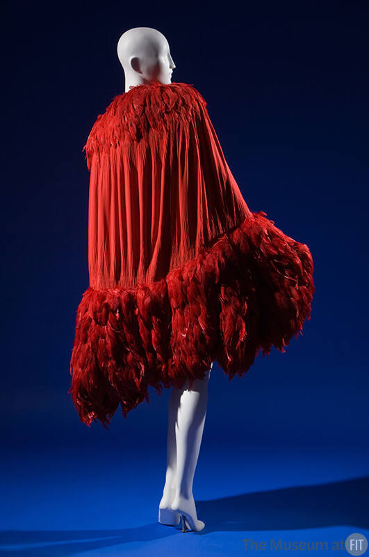Red evening cape smocked along curved edges of cocoon silhouette, crimson feather trim forming …