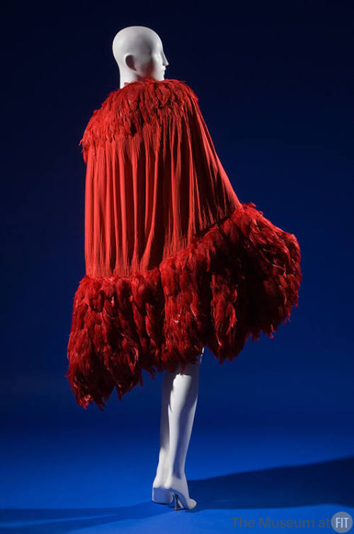 Red evening cape smocked along curved edges of cocoon silhouette, crimson feather trim forming …